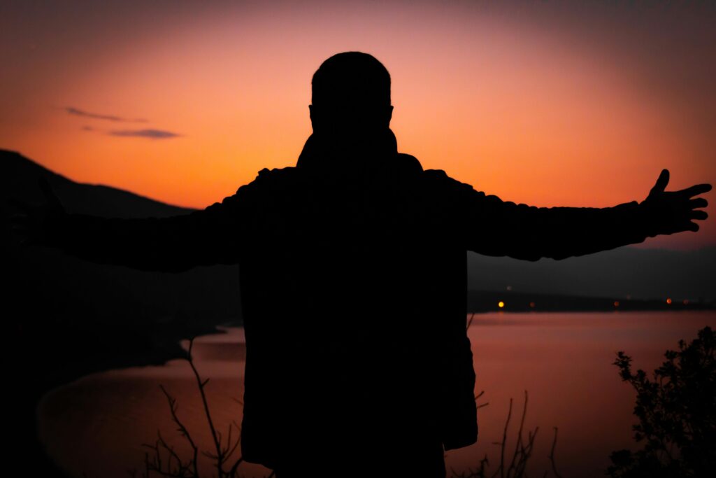 A person in silhouette with arms outstretched at sunset over a calm lake, creating a dramatic scene.